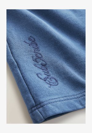 Mėlynos medvilnės audinys su tekstūruotu paviršiumi, palei kraštą siuvinėtu tamsiai mėlynu tekstu. Paprastas, be puošmenų dizainas.