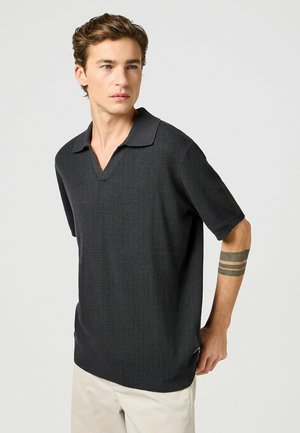 Ciemnoszara koszulka polo z krótkim rękawem, z głębokim dekoltem w kształcie litery V, teksturowana tkanina, oversize'owy krój i prążkowany dół. Model nosi jasne spodnie.