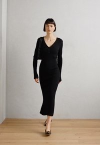 Abito a coste nero con maniche lunghe, scollatura a V e dettaglio con bottoni. Indossato con una borsa nera e tacchi lucidi marroni su un pavimento in legno.