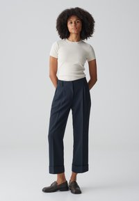 Helles beiges, geripptes Kurzarmoberteil, kombiniert mit dunkelblauen, hoch taillierten, gefalteten Hosen, die gerollte Ärmel und braune Slipper umfassen.