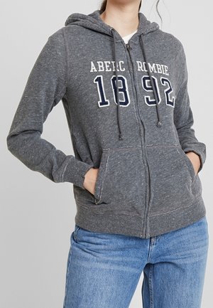 Personne portant un sweat à capuche gris zippé avec le texte "ABERCROMBIE 1892" et un jean bleu, mains dans les poches avant sur un fond uni.