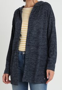 Marinblå, hoodad cardigan i mjukt, strukturerat tyg, med en lös passform och öppen framsida, bärs över en gulrandig skjorta.