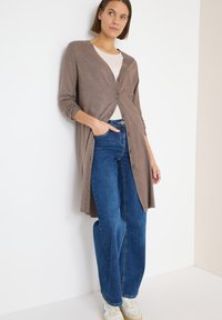 Lange, bruine cardigan met een V-hals, knoopsluiting aan de voorkant en lange mouwen, gecombineerd met blauwe flared jeans en witte sneakers. Glad aanvoelende stof.