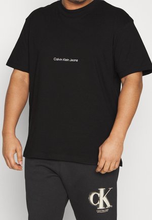 Uomo che indossa una t-shirt nera Calvin Klein Jeans e pantaloni neri con il logo bianco CK sulla coscia sinistra su sfondo bianco.