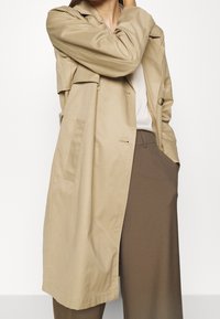 Beige pláštěnka s volným střihem, která má velké knoflíky, boční kapsy a hladkou texturu látky. Nosí se přes lehký top a hnědé kalhoty.