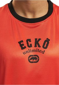 Rød kortærmet t-shirt med sort krave, har et fedt sort "ECKŌ unlimited" logo og et næsehornsgrafik i midten, glat stofstruktur.