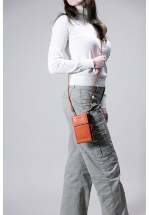 Orange leather crossbody bag with a smooth texture, featuring a front flap pocket, worn on light-grey, loose-fitting cargo pants.