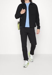 Czarna kurtka zapinana na guziki, jasnoniebieska koszula, czarne spodnie oraz szaro-zielone sneakersy z neonowymi akcentami. Kurtka posiada kieszeń na piersi i gładką fakturę.