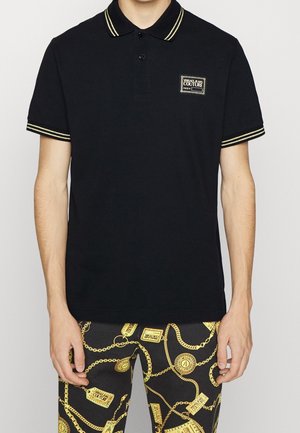 Schwarzes Polohemd mit kontrastierendem, gelb gestreiftem Kragen und Ärmeln, mit einem kleinen weißen Logopatch auf der Brust. Baumwollstoff.