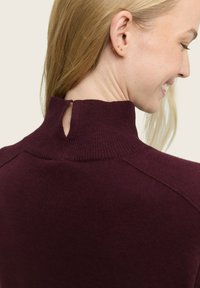Maglione a maglia borgogna con collo alto e dettaglio di taglio sul retro. Il tessuto è morbido con accenti a coste sul collo e sulle spalle.