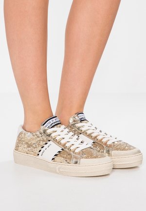 Füße, die goldene Pailletten-Low-Top-Sneakers mit weißen Schnürsenkeln und weißen Einsätzen tragen, stehen auf einer weißen Oberfläche.