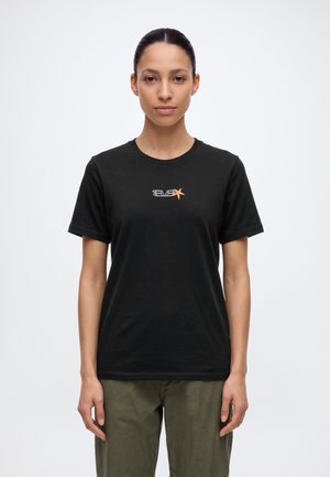 Une femme porte un T-shirt noir à manches courtes orné d’un petit logo sur la poitrine et un pantalon olive, sur un fond blanc uni.