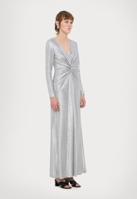 Ασημένιο φόρεμα με μακριά μανίκια, βαθύ V-λαιμόκοψη και κόμπο στη μέση; φτιαγμένο από γυαλιστερό, υφασματώδες υλικό. Μαύρα παπούτσια με ψηλό τακούνι.