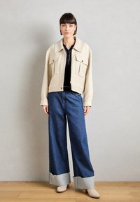 Beige cropped jakke med to frontlommer, kombinert med vide blå jeans med slitte sømmer og grå detaljer. Beige ankelstøvler fullfører antrekket.
