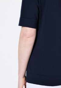 Navy modrá krátká pletená halenka s texturovaným povrchem, která má ohrnuté manžety a rovný spodní lem. Zadní pohled ukazuje hladký design.