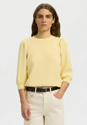 Kvinde med bølget brunt hår iført en blege gul top med puffede ærmer, der er stoppet ned i off-white bukser med et mørkebrunt bælte, stående med den ene hånd i lommen.