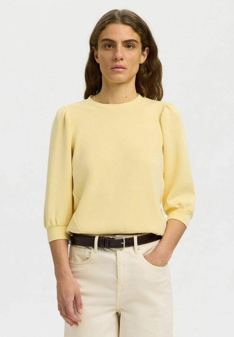 Femme aux cheveux bruns ondulés portant un haut jaune pâle à manches bouffantes rentré dans un pantalon écru avec une ceinture marron foncé, debout avec une main dans la poche.