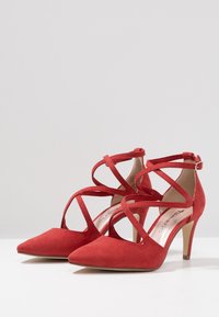 Chaussures à talons hauts en suède rouge avec un bout pointu, des brides entrecroisées et une boucle à la cheville. Le talon mesure environ 7 cm de hauteur.