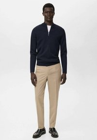 Suéter de cremallera azul marino sobre una camisa blanca, combinado con pantalones beige claros y zapatos negros. Textura suave, diseño ajustado, detalles mínimos.