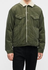 Personne portant une veste en velours côtelé vert olive avec un col en polaire blanche et une fermeture à boutons sur le devant, associée à un pantalon noir.