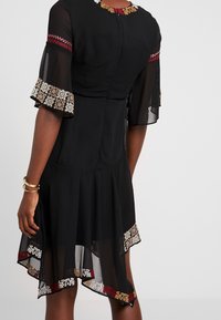 Musta mekko, jossa on läpinäkyvät hihat ja monimutkaiset kirjailut punaisella, kullalla ja valkoisella sekä levenevä helma, jossa on kontrastisia kuvioita.