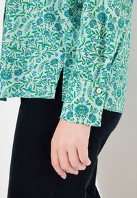 Camisa de manga larga con un patrón floral en verde claro que presenta flores azules y acentos de hojas, terminada con un puño abotonado y aberturas laterales.