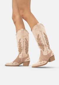 Nogi w beżowych zamszowych kowbojkach do połowy łydki z brązowymi aplikacjami w kształcie liści i niskimi, płaskimi obcasami, jedna stopa lekko uniesiona.