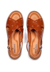 Sandalias de cuero marrón con diseño cruzado, tachuelas de metal, correa ajustable y plantilla acolchada. Suela plana y detalle de costura reforzada.
