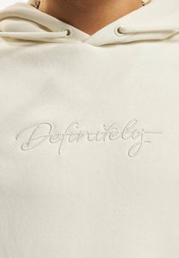 Light beige hoodie with a soft texture and embroidered cursive text "Definitely" in a slightly contrasting thread above the chest area.