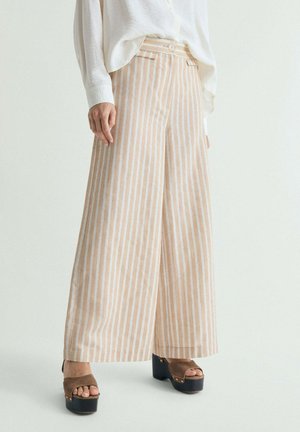 Persona con pantalones anchos de rayas verticales beige y blancas, camiseta blanca y sandalias plataforma marrones, de pie sobre un fondo sencillo.