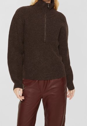 Femme portant un pull en maille côtelée marron foncé avec une fermeture éclair à l'avant et un pantalon en cuir bordeaux, debout devant un fond uni.