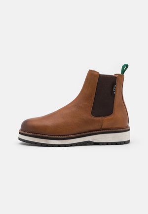 Botte Chelsea en cuir marron avec empiècement latéral élastique noir, semelle épaisse blanche et noire, et languette en tissu verte à l'arrière.