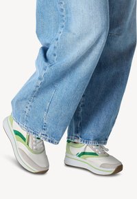 Sneaker da uomo bianco con dettagli verde, sovrapposizione testurizzata e particolari grigi. Abbinato a jeans a gamba larga blu chiaro.