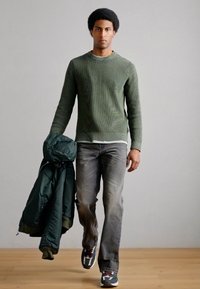 Zeleni rebrasti pulover, sive kavbojke, temno zelena jakna obešena čez ramo in vzorčaste superge. Gladek tekstura, oprijet kroj in priložnostni slog.