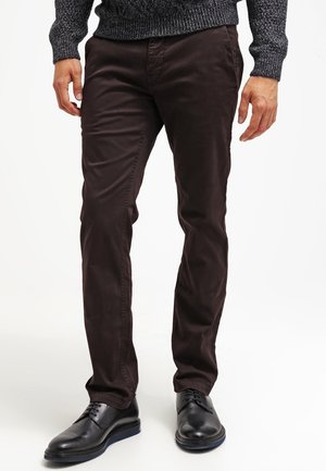 Hombre con pantalones ajustados de color marrón oscuro, zapatos de vestir negros de cuero y un suéter de punto gris oscuro, de pie frente a un fondo blanco.