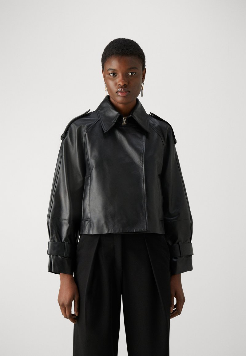 Μαύρο δερμάτινο cropped τζάκετ με μεγάλο γιακά, επι shoulder epaulets και ζώνες στους καρπούς. Λείο υφή, μινιμαλιστικός σχεδιασμός. Μαύρο παντελόνι από κάτω.