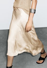 Jupe midi en satin beige avec un design fluide, une texture légère et un éclat subtil, associée à des sandales plates noires et un bracelet manchette en argent.