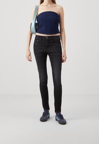 Joven con una blusa sin tirantes azul, jeans ajustados negros, zapatillas azules y llevando un bolso de hombro azul claro sobre un fondo liso.