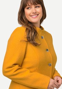 Cappotto di lana giallo con colletto alto, dotato di chiusura a doppio bottone e dettagli testurizzati sottili sul tessuto.
