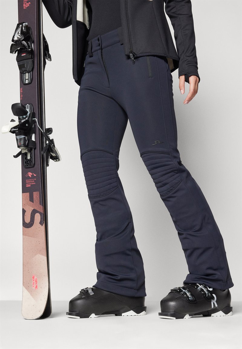 Marineblaue Skihosen mit gesteppter Kniefärbung, schlanker Schnitt und mit Reißverschlüssen versehenen Taschen, kombiniert mit schwarzen Skischuhen und einem pinken, gemusterten Snowboard.