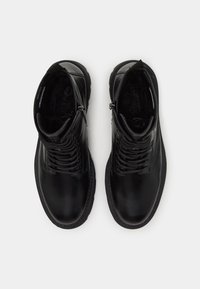 Des bottes de cheville en cuir noir présentent une surface lisse, un bout rond et un système de laçage à l'avant avec une fermeture éclair sur le côté. Semelle intérieure texturée visible.