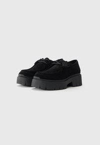 Chaussures en daim noir avec une semelle en caoutchouc épaisse, présentant un design cousu, un système de laçage et une étiquette ronde "HUGO" en accent.