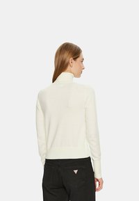 Pullover bianco con collo a imbuto, polsini e orlo a coste, texture morbida, design aderente; indossato con jeans neri con una patch logo sulla tasca posteriore.