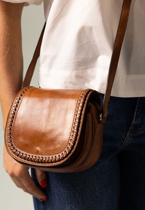 Sac bandoulière en cuir marron avec un rabat arrondi, des coutures complexes le long des bords et une sangle réglable. Surface texturée et design compact.