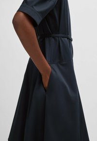 Navy blue dress with a fitted waist, short sleeves, and a flared skirt. Features side pockets and a smooth, matte texture.