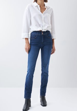 Vrouw met donkerblauwe, high-waist skinny jeans, een wit hemd met knopen dat in de taille is gestrikt, en zwarte enkellaarsjes met hak, staand tegen een effen achtergrond.