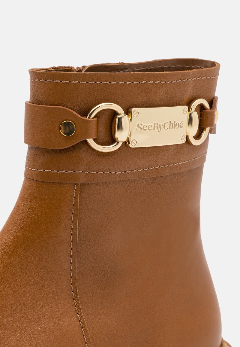 Bruine leren enkelboot met een gouden metalen kettingaccent en een plaatje met "SeeByChloé". Glad oppervlak en zichtbare stiksels.