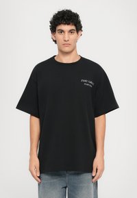 Svart bomulls oversized T-skjorte med korte ermer og rund hals, med hvit tekst på venstre side som leser "JUST CAVALI."