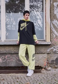 Mustard suur Nike t-särk kollase logoga, kihilise kollase kapuutsiga. Hele roheline spordipüksid sisaldavad sarnast logo, koos valgete tossudega.