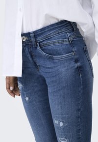 Jeans de mezclilla azul oscuro con áreas desgastadas, con un diseño ajustado, cinco bolsillos y costuras amarillas en contraste a lo largo de las costuras.